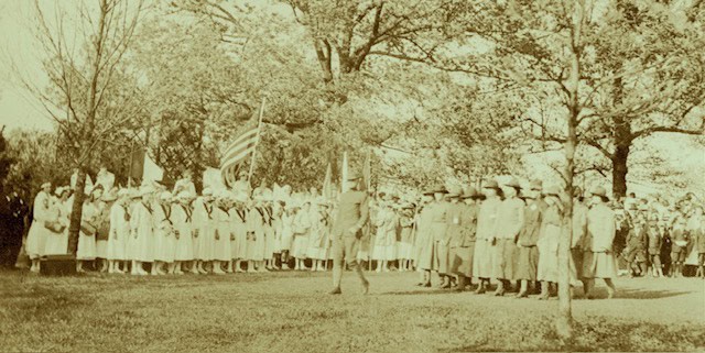 Sepia-toned image showing a small group of women on parade, led by a man in uniform. A large group of students with flags look on. They are on a grass lawn, surrounded by trees.