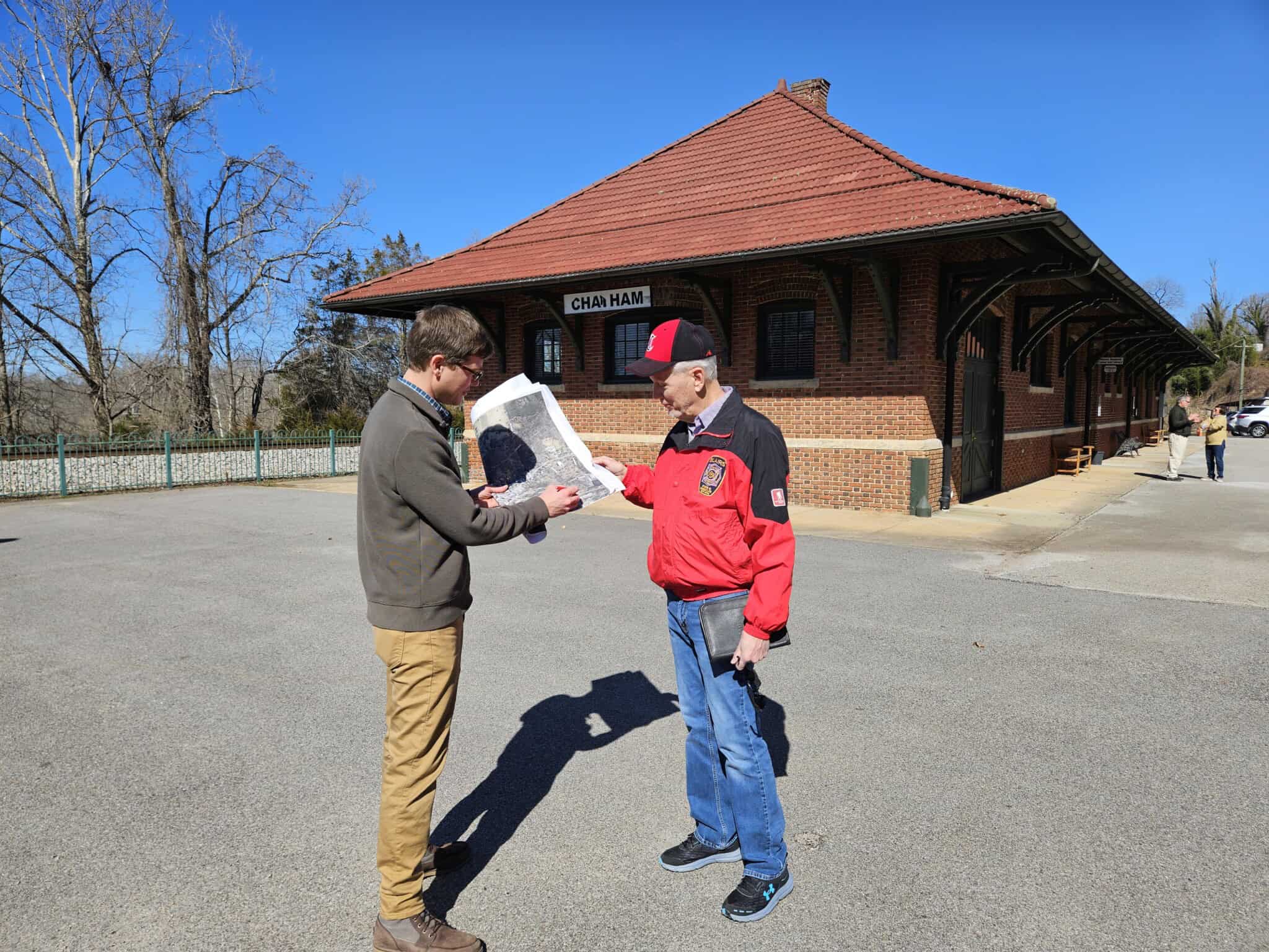 Two people stand outdoors, looking at a large aerial image of the town together. They're near the former train depot, a large one-story brick building. In the background, two other people talking, and railroad tracks are beyond a fence.