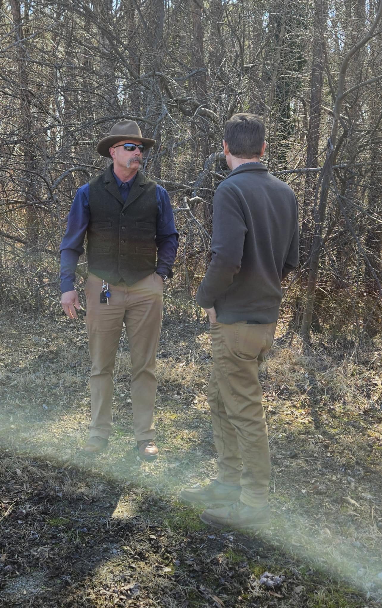 Two men standing and talking in front of a wooded area, both wearing casual outdoor attire including jackets and pants. One man wears a hat and sunglasses. Sparse undergrowth is visible around them.