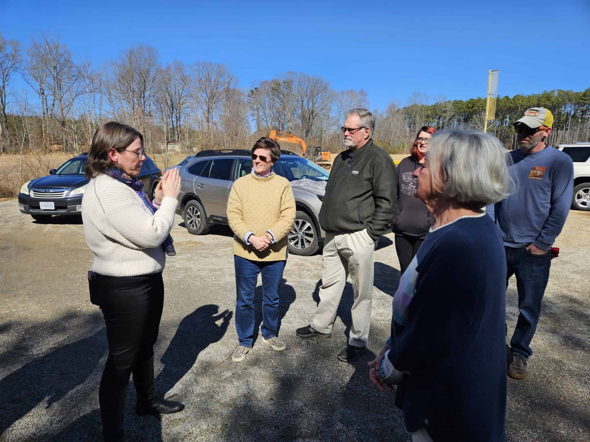 A group of people stands in a parking lot, engaging in conversation. It is a clear day. Cars, construction equipment, and trees are visible in the background.