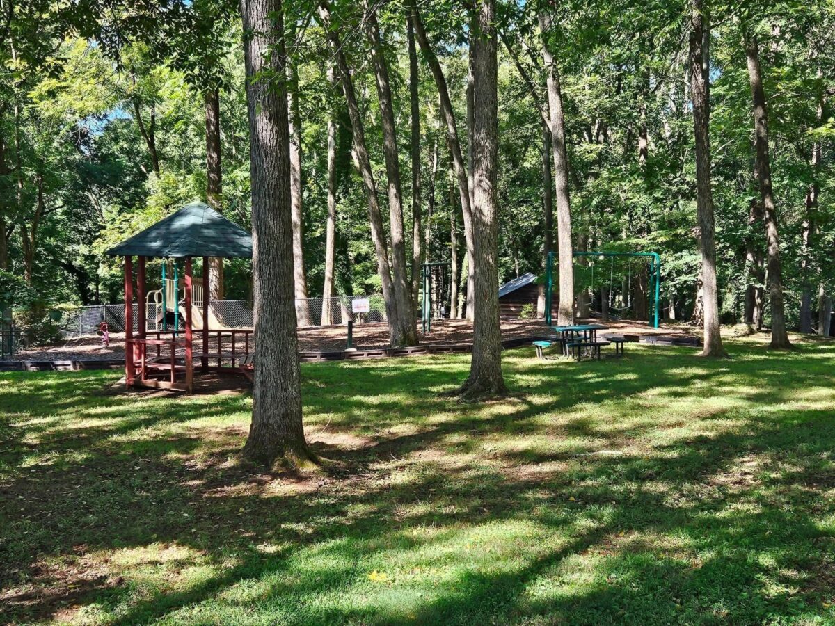 Nestled in a wooded area, this playground features a slide, swings, and a gazebo. Sunlight filters through the trees, casting playful shadows on the grass.