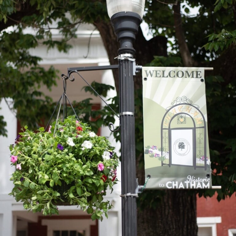 Street lamp adorned with a hanging flower basket and "Welcome to Historic Chatham" sign, with trees and the courthouse building painting the backdrop.