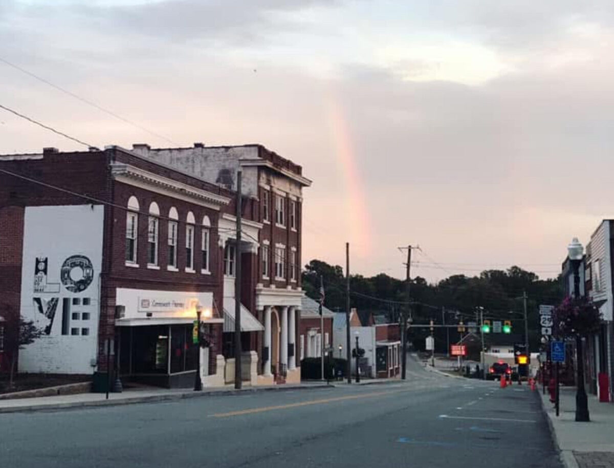 Chatham's Main Street is lined with brick buildings. A faint rainbow graces the cloudy sky, adding charm to the scene, while a vibrant "LOVE" mural brightens one wall.