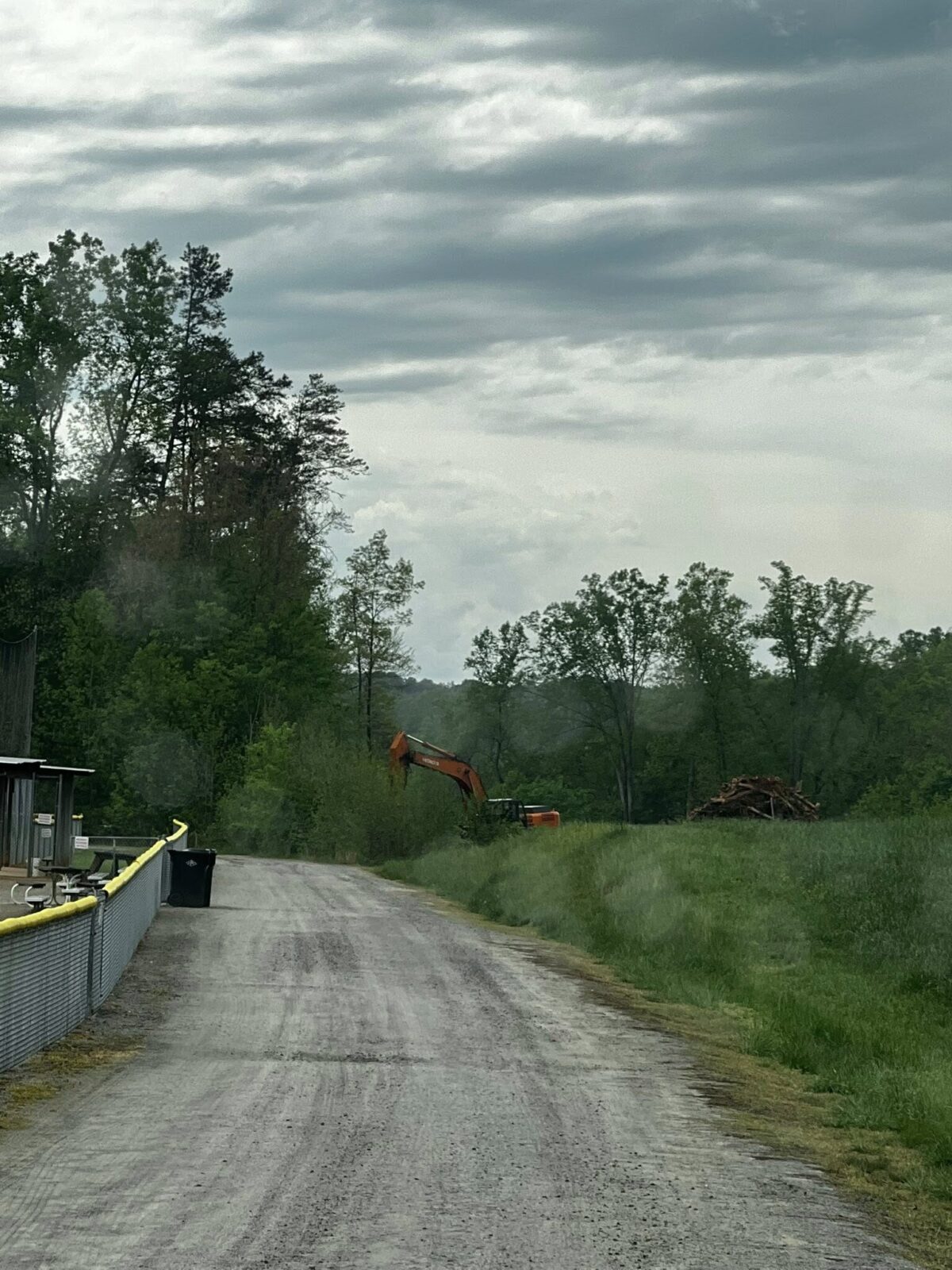An orange excavator is visible beyond the gravel lane beside the Harris baseball fields.