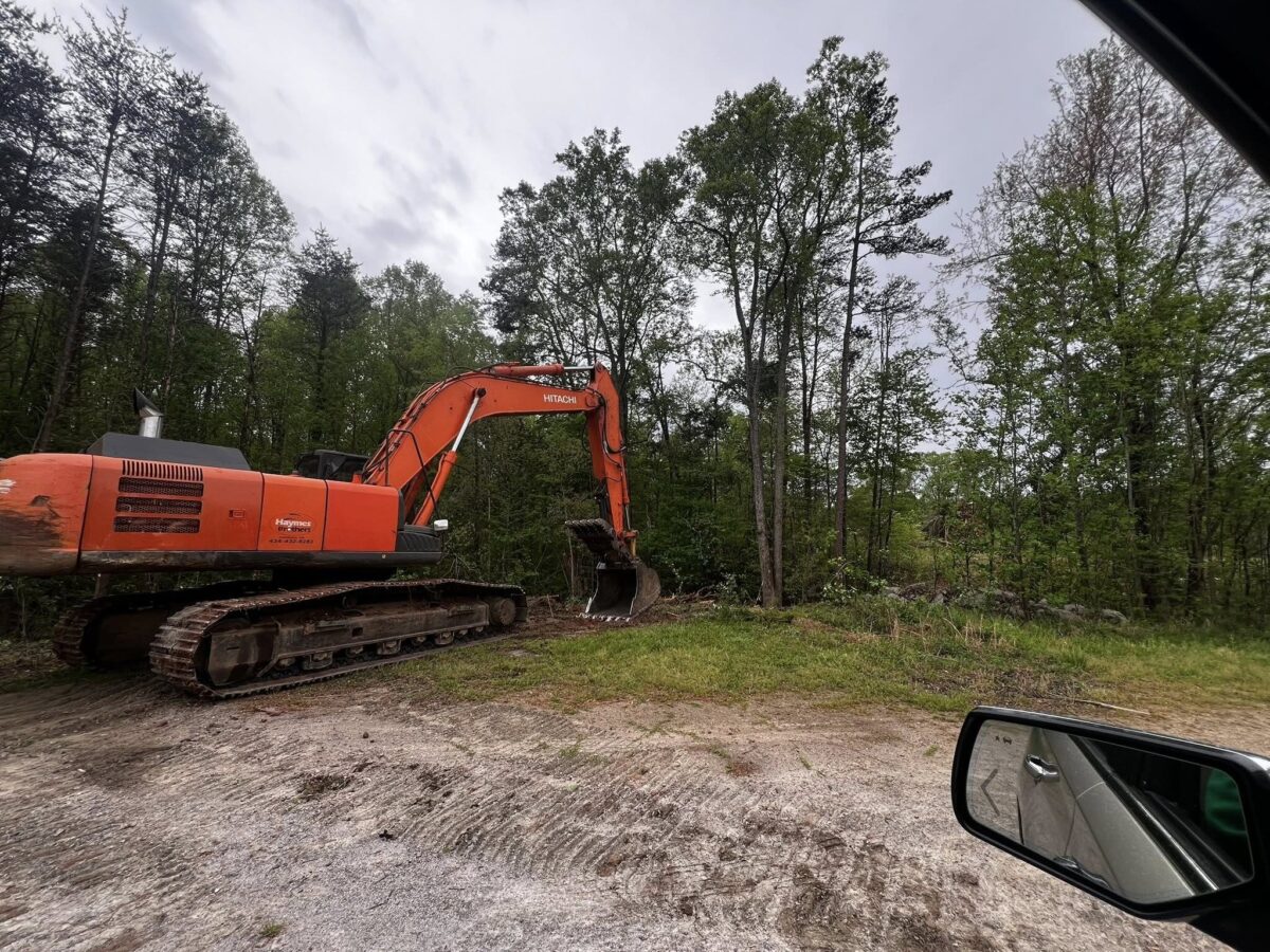 An orange excavator is working at the edge of a dirt clearing next to a wooded area under an overcast sky.