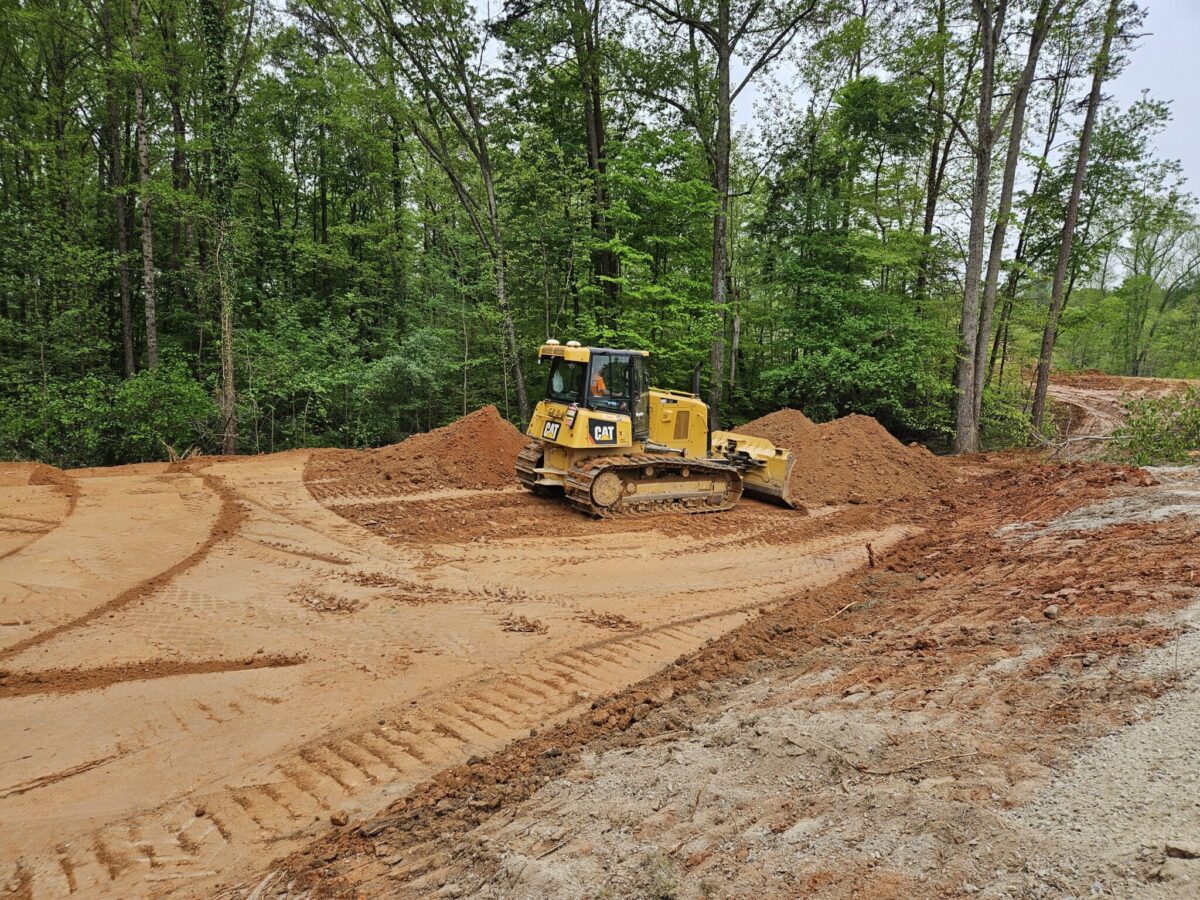A yellow bulldozer is leveling dirt at a site surrounded by trees, with tread tracks and earth piles visible in the area.