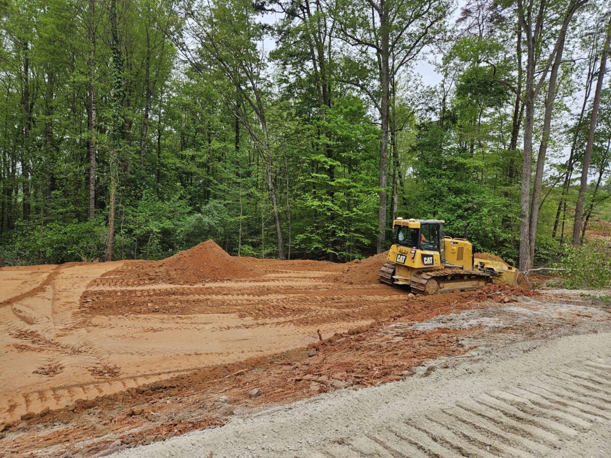 A yellow bulldozer is at the edge of a newly cleared area backed by trees, with piles of soil and tread tracks visible on the ground.