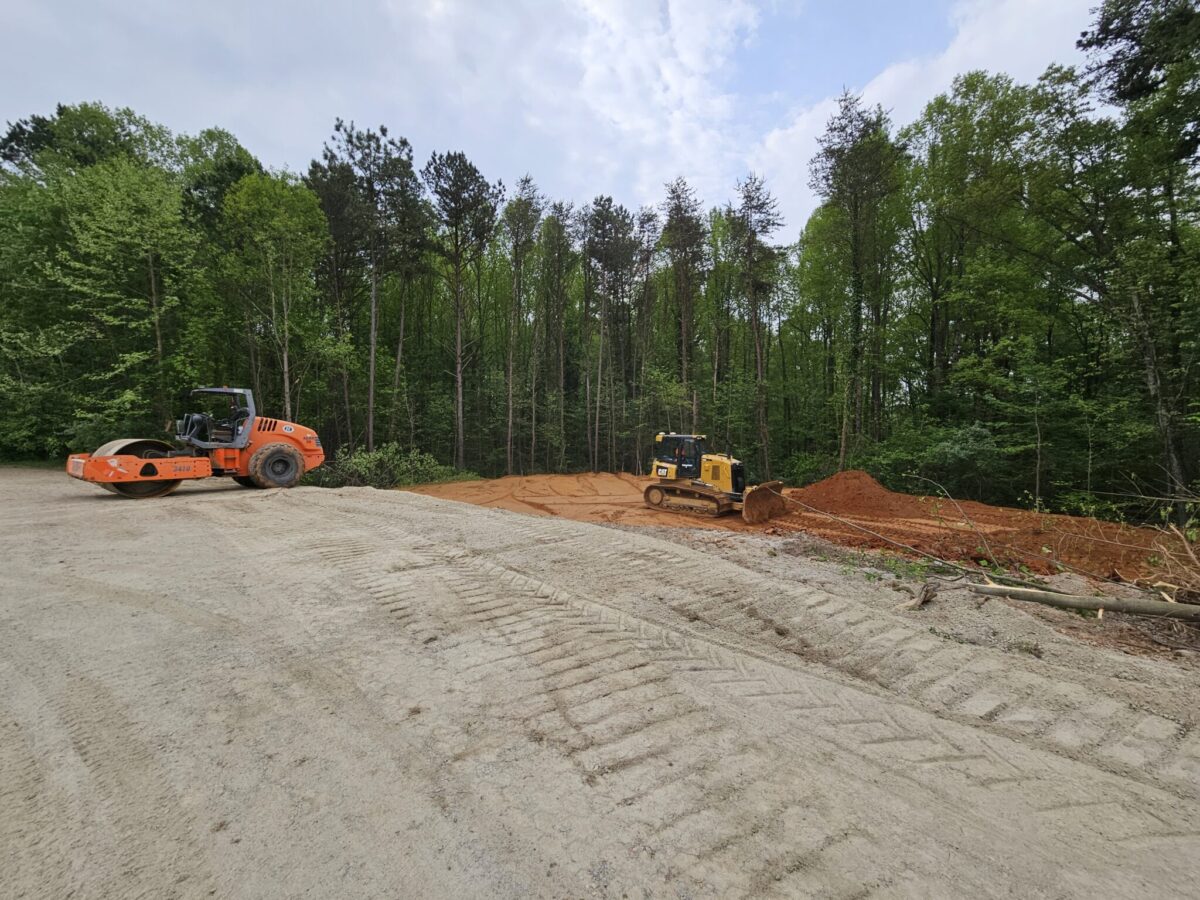 A construction site with a steamroller and a bulldozer moving dirt beside a wooded area.